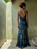 A woman with a red flower in her hair stands in a sunlit, earthy-toned corner, wearing the Rimmba Scarlette Silk Dress - Indigo Circle, featuring a low back and circular indigo patterns, facing away from the camera.