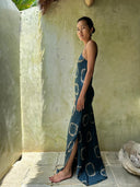 A woman stands barefoot on a tiled floor, wearing the Scarlette Silk Dress - Indigo Circle by Rimmba | Sustainable Clothing, in a sunlit corner with pale green textured walls and a large seashell nearby.