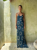 A woman stands against a light textured wall, wearing Rimmba's Scarlette Silk Dress - Indigo Circle, a hand-dyed sustainable long dark silk dress with indigo circle patterns. Her hair is tied back as she looks neutrally at the camera.