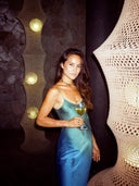 A woman in the Scarlette Silk Dress - Indigo Ombre by Rimmba | Sustainable Clothing holds a glass of white wine, standing before glowing sculptural woven lamps and a stone wall backdrop.