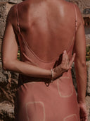 Wearing the Scarlette Silk Dress in pink circle motif by Rimmba | Sustainable Clothing, a person stands back to camera, one arm bent behind as sunlight highlights beaded jewelry and warm shadows on the hand-dyed silk.
