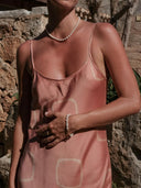 A woman in the Scarlette Silk Dress - Pink Circle Motif by Rimmba | Sustainable Clothing stands with one hand on her waist in sunlight, displaying tan lines; stone walls and greenery are in the background.
