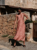A woman stands outdoors against a rustic stone wall, wearing the Scarlette Silk Dress - Pink Circle Motif by Rimmba | Sustainable Clothing, a sleeveless hand-dyed silk piece with her hand in her pocket, gazing downward.