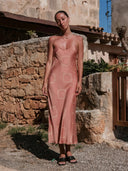 A woman stands outdoors in front of a stone wall, wearing the Scarlette Silk Dress - Pink Circle Motif by Rimmba | Sustainable Clothing, paired with black sandals and a pearl necklace in a sunny, rustic setting.