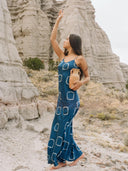A woman stands barefoot on rocky terrain, holding a tan jug and gracefully raising one arm, wearing the Scarlette Silk Dress - Indigo Circle by Rimmba | Sustainable Clothing against pale rock formations.