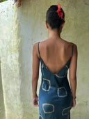 A woman with short dark hair wears the Scarlette Silk Dress - Indigo Circle by Rimmba | Sustainable Clothing, styled with thin straps, a red flower in her hair, and gold hoop earrings, standing before a textured light green wall.