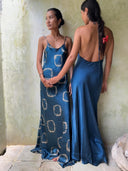 Two women indoors wear Rimmba's Scarlette Silk Dress - Indigo Circle. One faces forward, showing the tie-dye design and thin straps; the other shows off the low back, with a red bow adorning her braided hair.