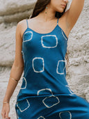 A woman wears the Rimmba Scarlette Silk Dress - Indigo Circle, featuring blue silk with circular patterns, standing against a textured, light stone wall. Only her torso and part of her face are shown, with her arm raised and hair over one shoulder.
