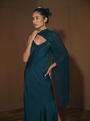 A woman in the Scarlette Silk Dress - Indigo by Rimmba | Sustainable Clothing, featuring an asymmetrical sheer cape, stands indoors against a brown background, gazing to the side with one hand on her neck and one on her hip.