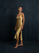 Left side view of woman wearing midi silk dress in mango colour with thin straps and slit against a dark background.
