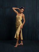 Left side view og woman wearing silk midi silk dress in mango colour with thin straps and slit against a dark background.