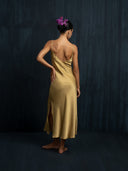 Barefoot woman facing a dark backgrund wearing luxury midi silk dress in mango colour with thin straps. Bun hairstyle with beautiful purple flower.