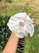A hand holds a large, white Silk Scrunchie by Rimmba | Sustainable Clothing outdoors, with green grass and leafy plants in the background—ideal for preventing hair damage.