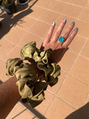 A hand with silver nails and a blue-green ring wears two large olive green Silk Scrunchies by Rimmba | Sustainable Clothing on the wrist, resting on a sunlit tiled floor.