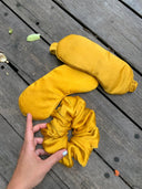 Two yellow Mulberry silk sleep masks and a Rimmba | Sustainable Clothing Silk Scrunchie rest on wood. A pale-nail-polished hand holds the scrunchie, ideal for preventing hair damage. Small leaves and debris are scattered nearby.