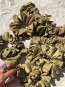 A hand with a blue oval ring reaches for Rimmba | Sustainable Clothing's large, olive green Silk Scrunchies, arranged on beige textured fabric and illuminated by sunlight, offering gentle hair protection.