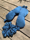 Two blue Rimmba | Sustainable Clothing mulberry silk eye masks and a matching Silk Scrunchies accessory rest on a wooden surface in sunlight, ideal for hair damage prevention.