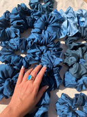 A hand wearing a blue gemstone ring touches a pile of Rimmba | Sustainable Clothing's Silk Scrunchies on a light surface with sunlight and soft shadows—ideal for hair damage prevention.