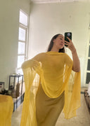 A person taking a mirror selfie indoors, wearing a long, flowing yellow dress with a silkchiffon yellow scarf draped gracefully over one arm. She is standing barefoot on a light-colored floor in a bright, minimalist room.