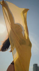 A woman standing  outdor wearring bikini, holding silkchiffon yellow scarf with two hands.