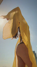 a woman photographed from behind wearing bikini, standing outdoors under a clear blue sky. A sheer, flowing yellow silkchiffon scarf is lifted by the wind, creating a soft, dramatic movement around their body.
