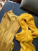 a mustard-yellow satin slip dress laid out on a dark blue sofa. Next to it is a matching sheer yellow silkchiffon scarf with a light, crinkled texture, loosely draped beside the dress.