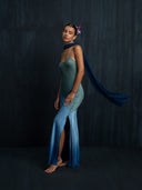 A woman stands barefoot against a dark backdrop wearing Rimmba’s Scarlette Silk Dress in Indigo Ombre, featuring a sleeveless design and thigh-high slit. A flowing blue scarf trails behind her, with pink flowers adorning her hair.