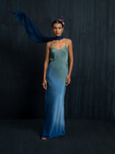 A woman stands against a dark background wearing the Scarlette Silk Dress - Indigo Ombre from Rimmba | Sustainable Clothing, paired with a flowing blue scarf and a pink flower in her hair. She faces forward with a calm expression.
