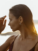 A woman with wet hair, wearing Rimmba's Silver Baby Hoop earrings in a hammered beaten design, stands outdoors in profile. Sunlight glows behind her, highlighting water droplets on her skin as she touches her forehead.