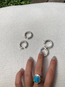 A hand with light blue nails and a large oval blue-green stone ring rests on fabric beside three pairs of Silver Baby Hoop earrings, hammered beaten design by Rimmba | Sustainable Clothing. Grass and wood are visible in the background.