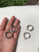 A hand shows two Rimmba | Sustainable Clothing Silver Baby Hoop earrings with a hammered beaten design, while another matching pair lies on textured fabric. Grass and wood can be seen in the background.