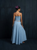 Woman wearing a baby indigo cotton strapless top and maxi cotton wrap skirt is facing backwards to a dark background.