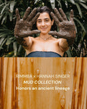 a woman standing outdoors in front of lush green foliage. She is smiling gently and holding both hands up toward the camera, palms facing out, showing that they are fully covered in dark mud. She is wearing a strapless, dark blue top and a simple necklace.