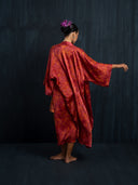 Barefoot woman is facing a dark background wearing flowy Mulberry silk kimono Red ecoprint. Hancrafted botanical-dyed kimono for sustainable luxury fashion and resort wear.

