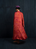 A flowing Mulberry silk kimono red ecoprint handcrafted botanical dyed. Wearing by barefoot woman is facing a dark background, bun hairstyle with purple flower.