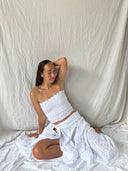 A woman wearing the Rimmba | Sustainable Clothing White Cotton Dayu - Elastic Bandeau Top sits on the floor in a long, natural cotton skirt, smiling gently against a white fabric backdrop.