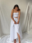 A woman wearing the White Cotton Embroidered - Suzzi's Skirt by Rimmba | Sustainable Clothing stands barefoot on white fabric, looking down and adjusting her maxi-length skirt against a backdrop of soft, light-colored drapes.
