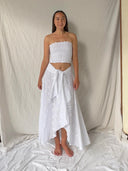A woman stands barefoot on a white sheet, wearing Rimmba's Suzzi's Skirt—a white cotton embroidered maxi skirt with a front tie, paired with a strapless top. Delicate floral embroidery stands out against the light beige backdrop behind her.
