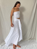 A woman stands barefoot on white fabric, wearing Rimmba | Sustainable Clothing's White Cotton Embroidered - Suzzi's Skirt. The long, flowy skirt features flower embroidery and a sash, set against a neutral draped backdrop.
