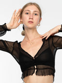 A young woman wearing gold jewelry poses with one hand raised, dressed in the Rimmba The Brand Zena Silk Chiffon Top - Black, a sheer 100% silk chiffon long-sleeve layered over a black bralette against a plain white background.