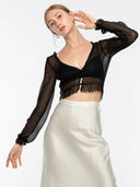 A woman with light hair pulled back poses in the Zena Silk Chiffon Top – Black by Rimmba The Brand, layered over a black bra and paired with a high-waisted ivory satin skirt. She accessorizes with hoop earrings against a plain background.