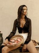 A woman with wet hair wears the Rimmba The Brand Zena Silk Chiffon Top in Black and white bottoms as she sits on a bench, holding a bamboo tray with red fish, looking to the side against a plain, light background.