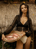 A woman in a sheer black Zena Silk Chiffon Top by Rimmba The Brand and white bottoms stands before a thatched hut, holding a basket of fresh fish and gazing off-camera with a relaxed expression.