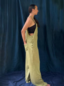 A woman stands in profile before a dark blue backdrop, wearing Rimmba | Sustainable Clothing's Zion Silk Overalls in Olive Green over a black sleeveless crop top, her hands casually tucked into the pockets.