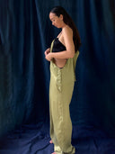 A woman with long dark hair stands sideways against a dark blue fabric backdrop, wearing a black top and loose olive green Zion Silk Overalls from Rimmba | Sustainable Clothing, styled in vintage 90s fashion as she looks down adjusting her outfit.