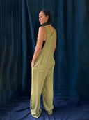 A woman stands sideways with her hands in the pockets of the Zion Silk Overalls in olive green by Rimmba | Sustainable Clothing, worn over a black top and posed against a dark blue fabric backdrop.