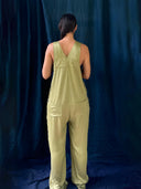 A person with dark braided hair faces away from the camera, wearing Rimmba | Sustainable Clothing’s Zion Silk Overalls in Olive Green, set against a dark blue fabric backdrop.