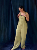 A woman with long dark hair stands barefoot against a dark blue fabric backdrop, wearing Rimmba | Sustainable Clothing’s Zion Silk Overalls in Olive Green over a black top. She looks down, gently holding one strap and smiling softly.