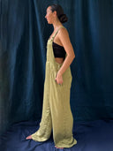 A woman stands in profile against a dark blue fabric backdrop, wearing Rimmba's Zion Silk Overalls in Olive Green over a black bandeau top. She is barefoot with her hair in a low bun and hands in pockets for a vintage 90s vibe.