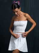 Woman wearing a white strapless top and mini wrap skirt against a dark background.